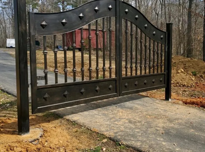 The Mayeaux Gate hung on square posts at the head of a new concrete apron, seen from the side of the drive with fresh earth banked either side and a red barn among bare trees behind.