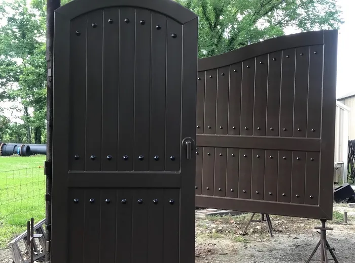 A tall arch-topped single leaf with a lever handle hanging on its post in the foreground, a wide single leaf on trestles behind it, both dark brown with black studs.