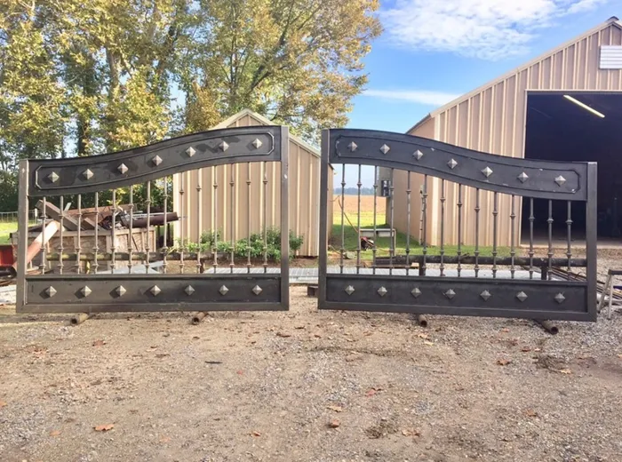 Both leaves standing on stands in the shop yard, still in bare grey steel, the S-curve rails and rows of diamond studs bright in the sun outside a tan metal workshop.