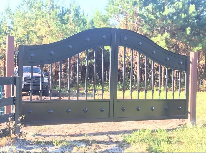 The Mayeaux Gate closed square-on between posts at the edge of a pine stand, its black leaves showing the S-curve top rail, turned balusters and studded bottom panel.