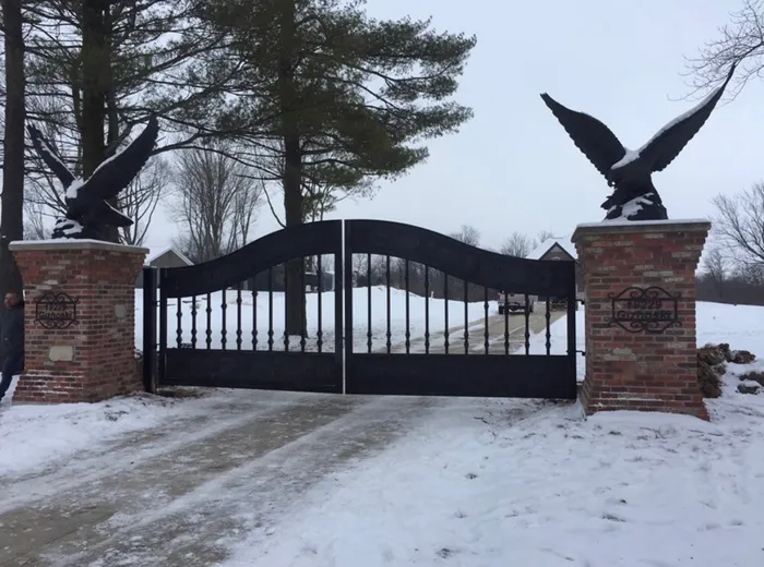 The design built again at another property, hung between brick piers topped with cast eagles and closed across a snow-covered drive, a scrolled iron name-and-number plaque set into each pier.