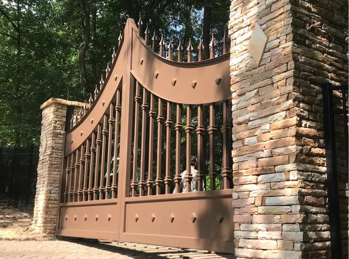 The gate in a light brown finish seen from the drive, spear finials rising along the swept rail between stacked-stone columns, a figure visible through the balusters.