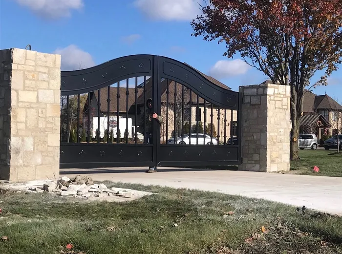 The design hung between pale stone columns on a bright autumn day, both leaves closed across a concrete drive, with a man in a dark hooded jacket standing behind the ironwork and a house and turning trees beyond.