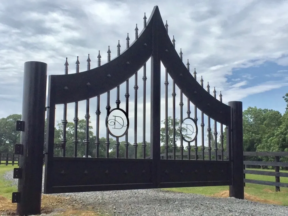 The Disotell Gate in a black finish closed across a gravel drive, its arched rails sweeping up to a peak at the centre under spear-point finials, an oval medallion carrying a script monogram set into each leaf, with a dark board fence and pasture beyond.