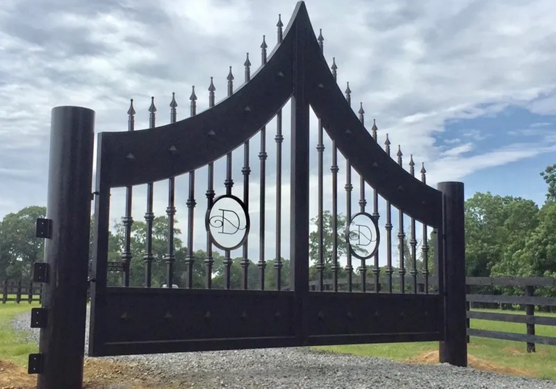 The Disotell Gate in a black finish closed across a gravel drive, its arched rails sweeping up to a peak at the centre under spear-point finials, an oval medallion carrying a script monogram set into each leaf, with a dark board fence and pasture beyond.