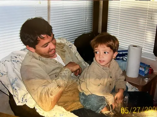 A photograph of an older print: Jason Satterley sitting with a young Jon, about three years old, both of them in matching tan work shirts covered in dirt from the shop.