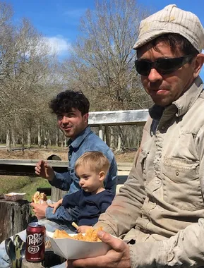 Jason in a weathered work cap and sun-faded shirt holding a basket of food outdoors, with Jon beside him in a denim jacket and Jon's young son sitting between them.